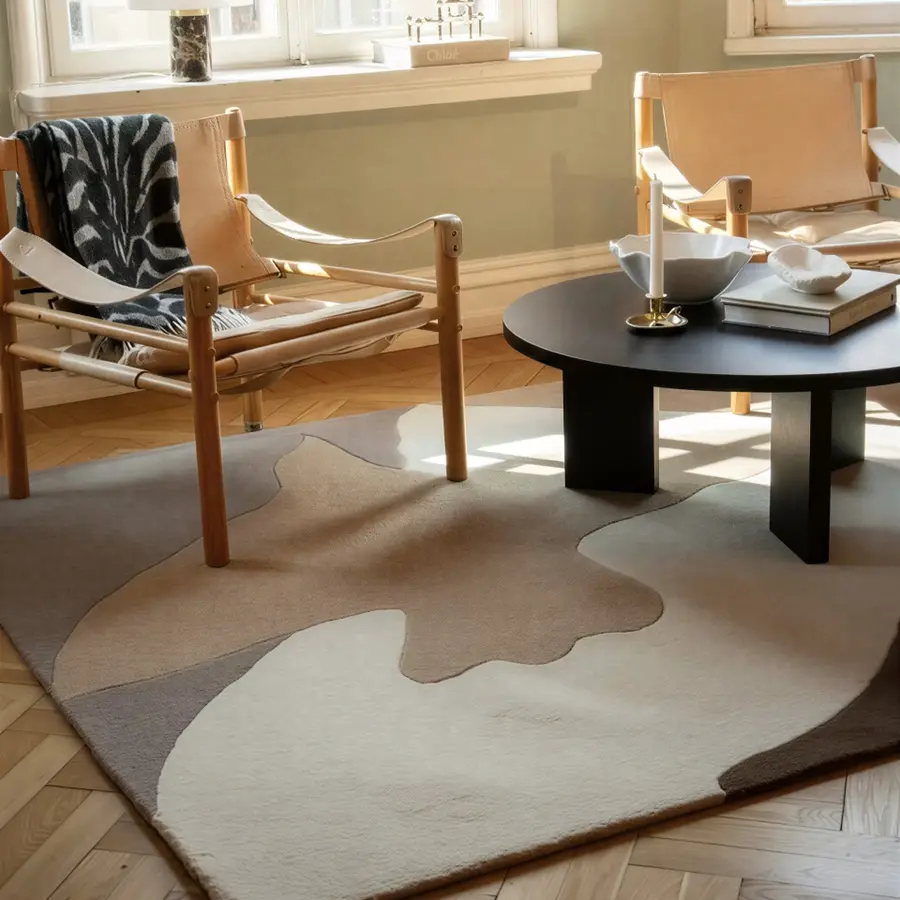 Two wooden chairs with tan leather seats, a black round coffee table, and a patterned rug in a sunlit room with light walls and parquet flooring.