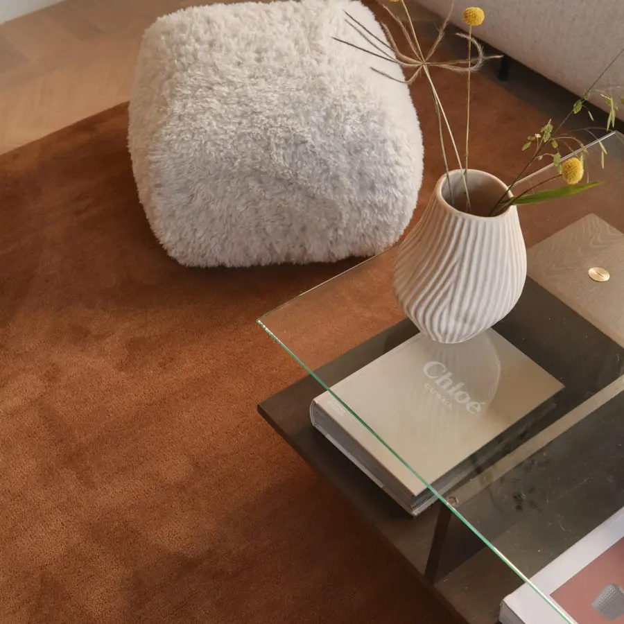A cozy living room setting with a fluffy pouf, a glass table holding a ribbed vase with flowers, and a book titled "Chloe" on a brown rug.