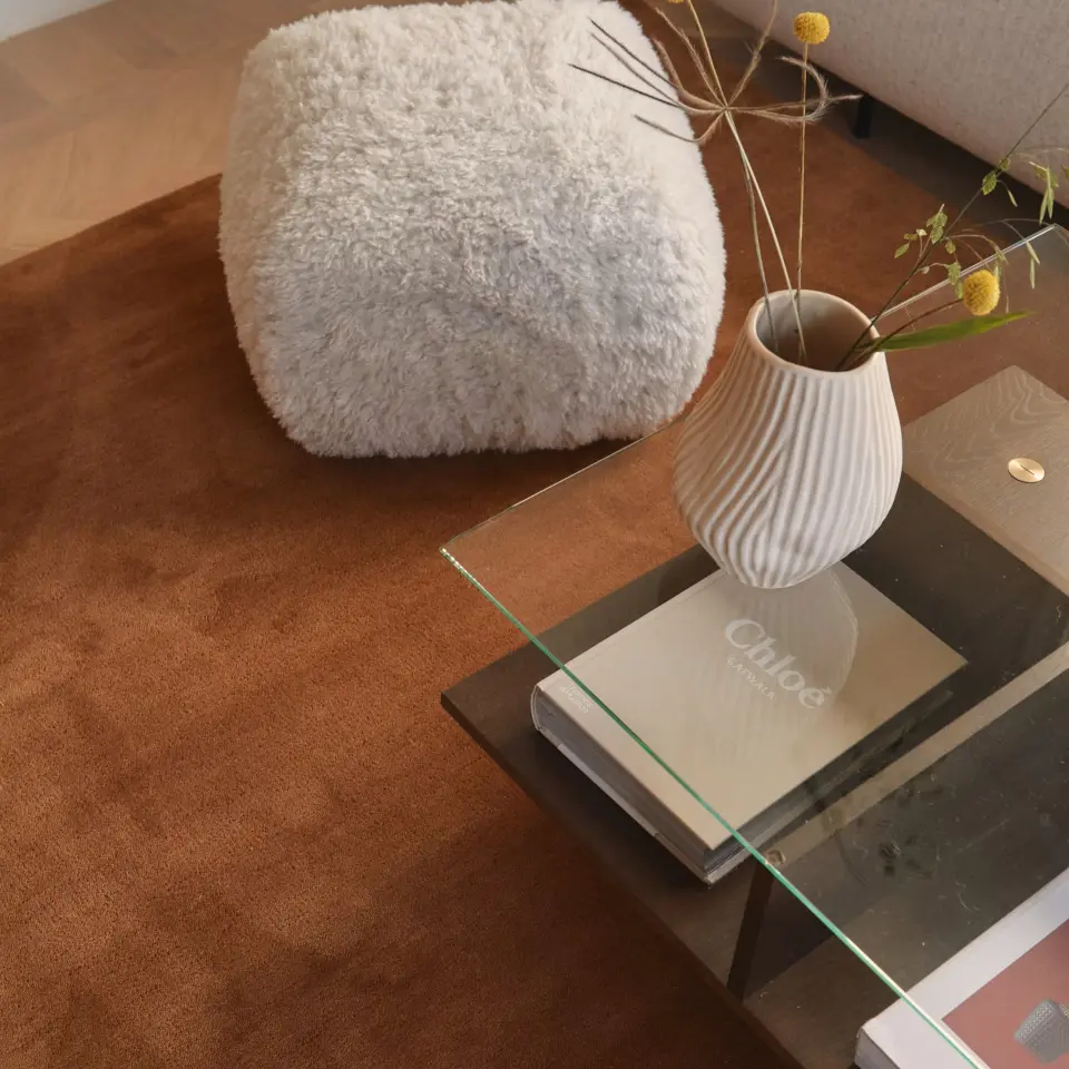 A cozy living room setting with a fluffy pouf, a glass table holding a ribbed vase with flowers, and a book titled "Chloe" on a brown rug.