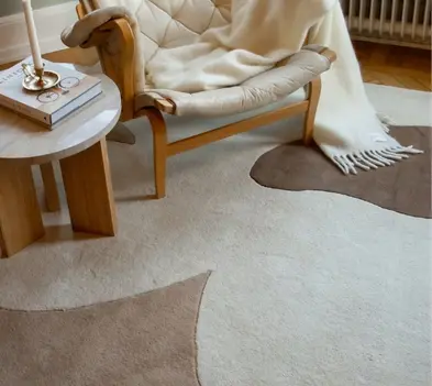 Cozy corner with a cushioned chair draped in a white blanket, beside a small wooden table with books and a candle on a patterned rug.